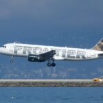 A Frontier Airlines passenger jet in mid-landing descends toward a runway beside a body of water.