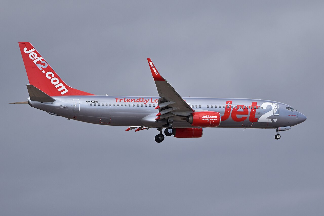 A silver Jet2 passenger airplane is captured mid-flight against a plain gray sky.