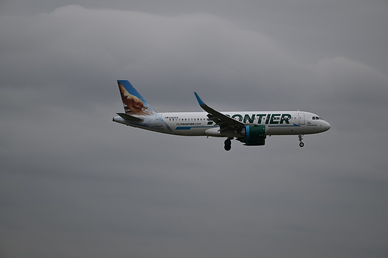 A Frontier Airlines jet flies through a gray, overcast sky with its landing gear extended, preparing to land.