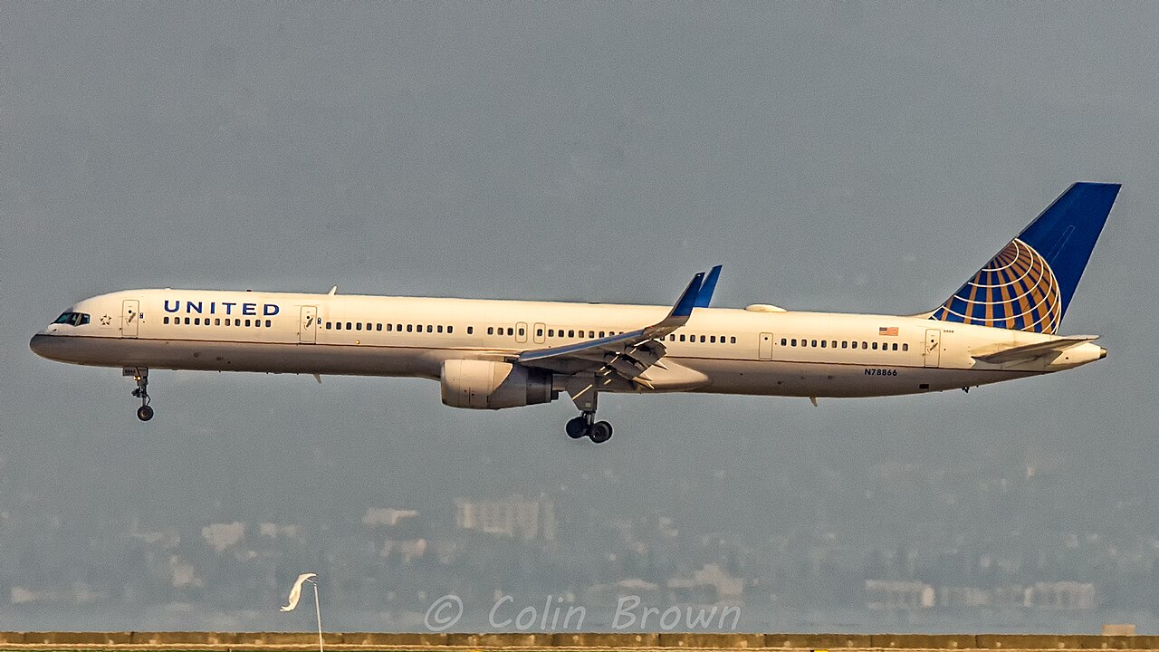 A long, narrow United Airlines jet with a white fuselage and “UNITED” written in blue letters flies low with its landing gear extended, preparing to land.