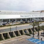 A wide view of a modern airport terminal labeled “Aeropuerto de Palma de Mallorca,” featuring a long white facade topped with a curved, lattice-like roof.