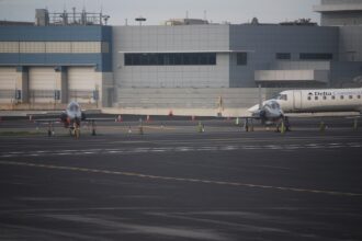 A wide airport tarmac scene with two small, sleek jet aircraft parked side by side in the foreground, each secured with orange cones and wheel chocks.