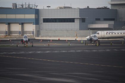 A wide airport tarmac scene with two small, sleek jet aircraft parked side by side in the foreground, each secured with orange cones and wheel chocks.