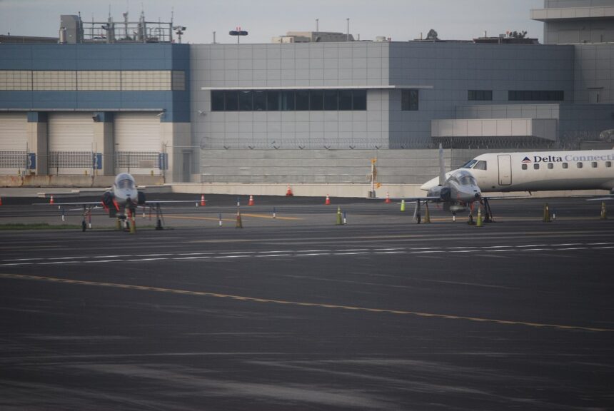 A wide airport tarmac scene with two small, sleek jet aircraft parked side by side in the foreground, each secured with orange cones and wheel chocks.