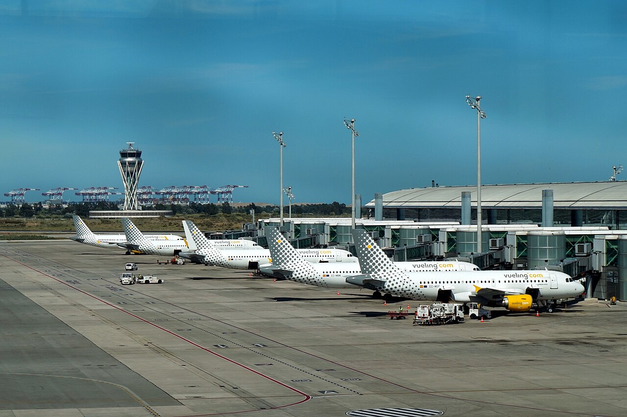 A row of Vueling Airlines planes is parked at airport gates along a modern terminal, each aircraft aligned nose-to-tail with jet bridges connected.