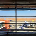 View from inside a terminal building looking out through large floor-to-ceiling windows onto an airport apron, where two IndiGo aircraft are parked under a clear blue sky with an expansive red-earthed runway in the background.