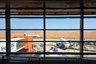 View from inside a terminal building looking out through large floor-to-ceiling windows onto an airport apron, where two IndiGo aircraft are parked under a clear blue sky with an expansive red-earthed runway in the background.