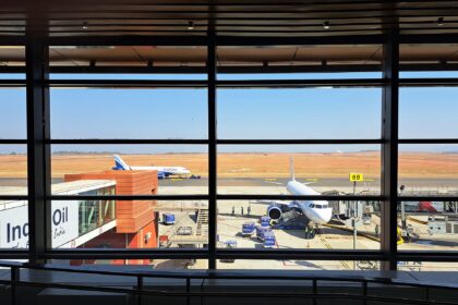 View from inside a terminal building looking out through large floor-to-ceiling windows onto an airport apron, where two IndiGo aircraft are parked under a clear blue sky with an expansive red-earthed runway in the background.