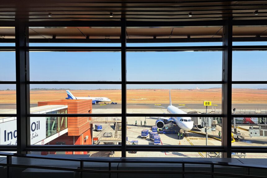 View from inside a terminal building looking out through large floor-to-ceiling windows onto an airport apron, where two IndiGo aircraft are parked under a clear blue sky with an expansive red-earthed runway in the background.