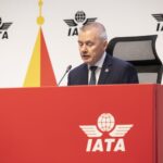 Willie Walsh, IATA's Director General sat on a table with a microphone. The IATA logo can be seen on both the red table and on the board behind him.