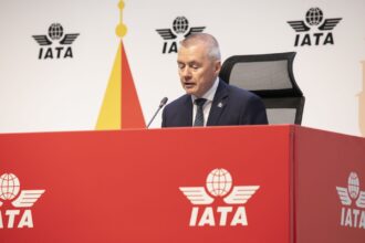 Willie Walsh, IATA's Director General sat on a table with a microphone. The IATA logo can be seen on both the red table and on the board behind him.