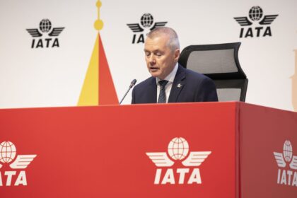 Willie Walsh, IATA's Director General sat on a table with a microphone. The IATA logo can be seen on both the red table and on the board behind him.