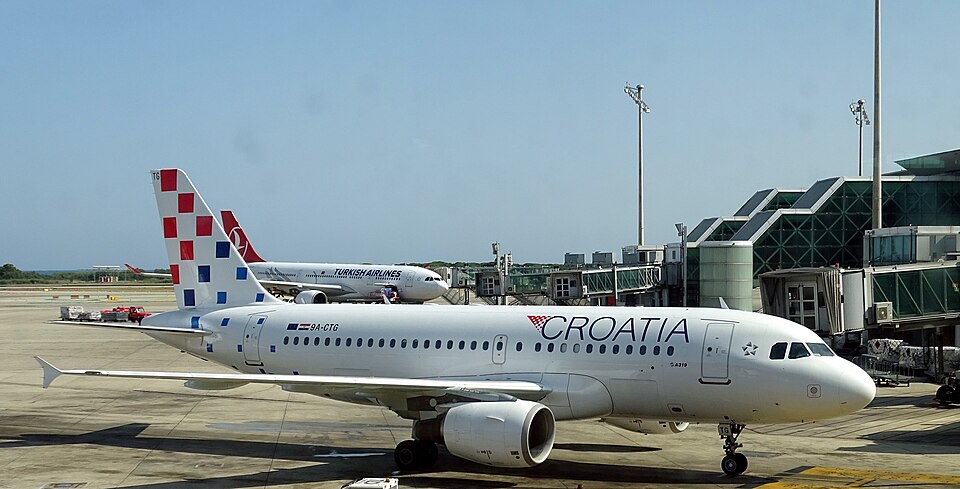 A high-angle, wide shot captures a Croatia Airlines Airbus A319-112 (registration 9A-CTG) parked at an airport terminal gate under a clear, bright sky.