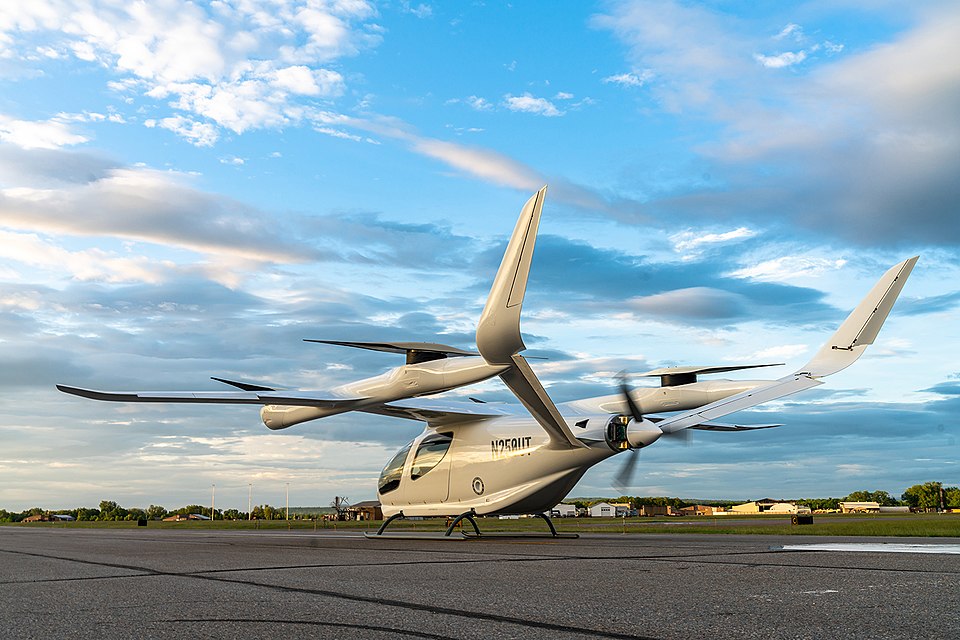 An all-electric ALIA-250 vertical takeoff and landing (eVTOL) aircraft by BETA Technologies, featuring a sleek white body, stationary on an airport tarmac under a blue, cloud-filled sky.
