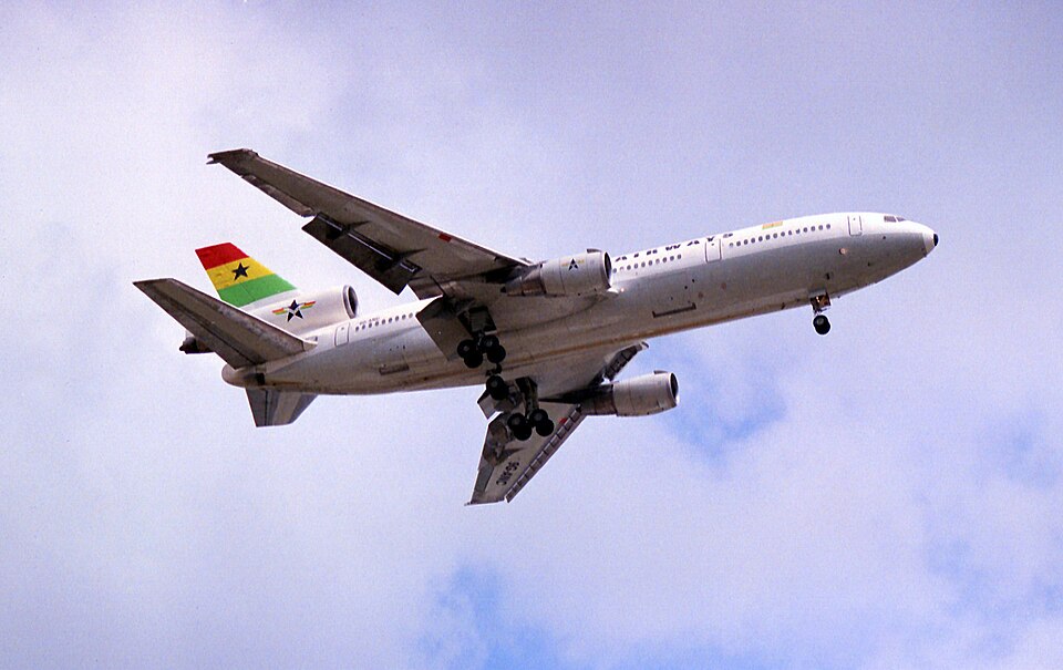 A low-angle shot of a Ghana Airways McDonnell Douglas DC-10-30 jet aircraft climbing into a pale blue, cloudy sky.