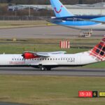 A Loganair ATR 72-600 turboprop aircraft, featuring its signature red-and-black tartan tail, taxies along an airport runway. In the background, the blue tail of a TUI Boeing 787 Dreamliner is visible