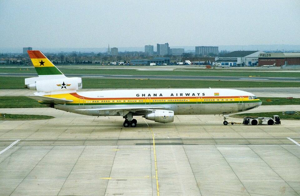 A side-profile view of a white Ghana Airways McDonnell Douglas DC-10-30 wide-body jet aircraft, featuring red, yellow, and green horizontal stripes along the body and the Ghanaian flag with its iconic black star on the tail fin.
