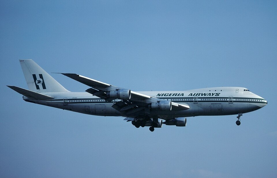 A side-profile image of a wide-body Nigeria Airways Boeing 747-200 aircraft, featuring the classic "Skypower" livery and logo, mid-flight against a clear, solid blue sky.