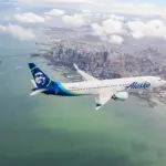 Alaska Airlines aircraft flying over a large blue body of water towards the skyscrapers in the distance.
