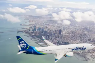 Alaska Airlines aircraft flying over a large blue body of water towards the skyscrapers in the distance.