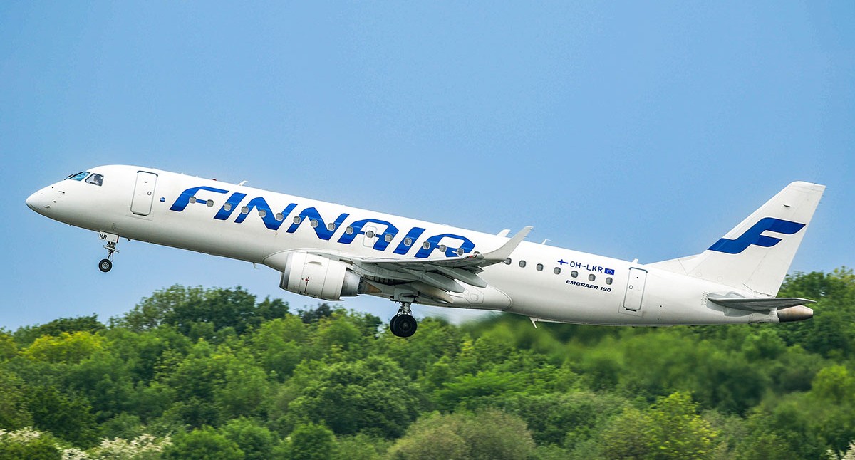 Finnair Embraer aircraft, white and blue livery, taking off, trees and blue sky in the background.