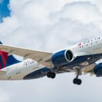 A Delta Air Lines Airbus A220-100 airplane with registration N101DU is shown in flight against a cloudy blue sky. The aircraft has its landing gear extended, and the Delta logo is visible on the body and tail