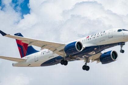 A Delta Air Lines Airbus A220-100 airplane with registration N101DU is shown in flight against a cloudy blue sky. The aircraft has its landing gear extended, and the Delta logo is visible on the body and tail