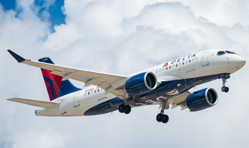 A Delta Air Lines Airbus A220-100 airplane with registration N101DU is shown in flight against a cloudy blue sky. The aircraft has its landing gear extended, and the Delta logo is visible on the body and tail