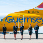 An image of a yellow and white aircraft on the ground with the word Guernsey on it and flight attendants standing in front of it.