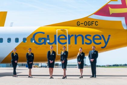 An image of a yellow and white aircraft on the ground with the word Guernsey on it and flight attendants standing in front of it.