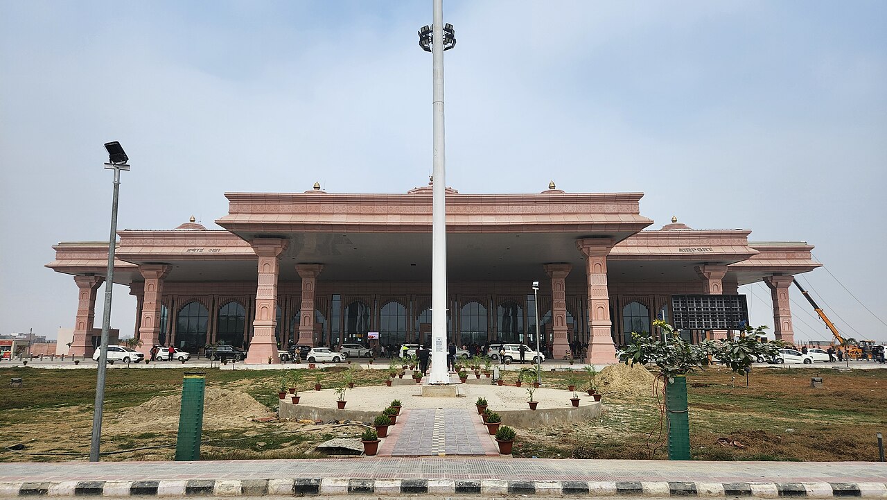 The façade of Ayodhya International Airport terminal building, designed in a traditional Indian temple architectural style with pink sandstone-coloured columns, ornate detailing, and arched entrances, set against a hazy sky.