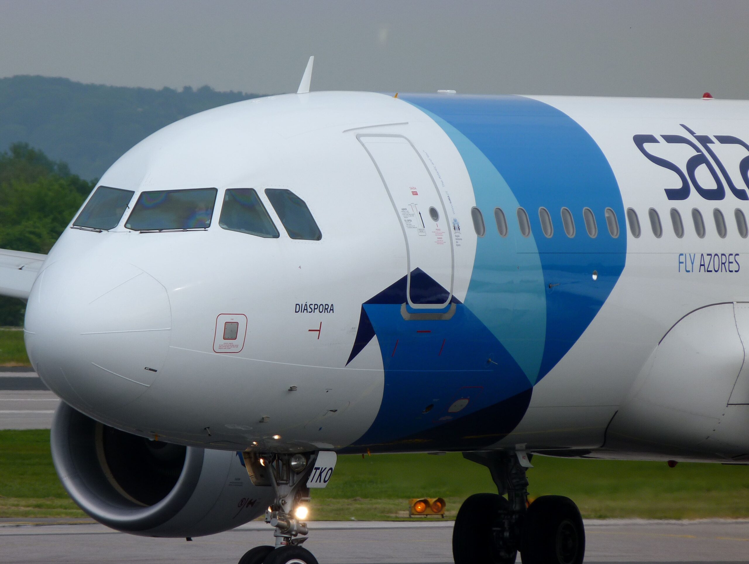 Close-up of a SATA Azores Airlines Airbus A320 on a runway, showing the blue-and-white nose section with cockpit windows, front landing gear and one engine intake visible, airline branding and “Fly Azores” text on the fuselage, with grassy surroundings and low, green hills beneath an overcast sky in the background.