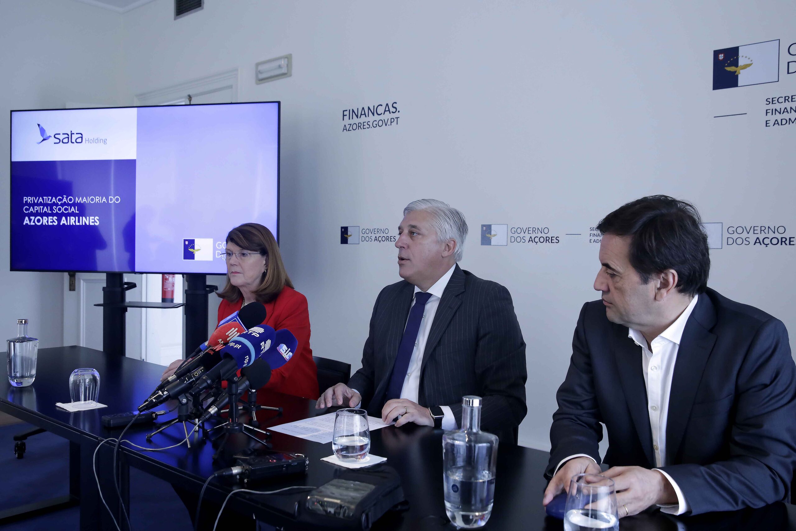 A panel of three officials sits at a long table during a press conference, with microphones and glasses of water in front of them. Behind them is a backdrop featuring the logo and text of the Government of the Azores’ Regional Secretariat for Finance, Planning and Public Administration. To the right, a large screen displays a presentation slide with the SATA Holding logo and the title “Privatização Maioria do Capital Social Azores Airlines,” indicating an announcement about the majority privatisation of Azores Airlines.