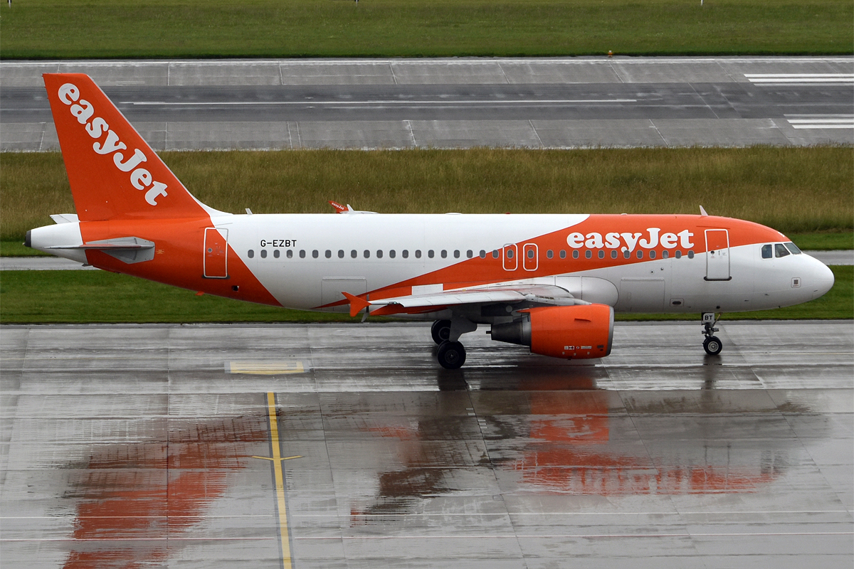 A bright orange-and-white easyJet Airbus A319 taxis along a wet airport apron on a rainy day.