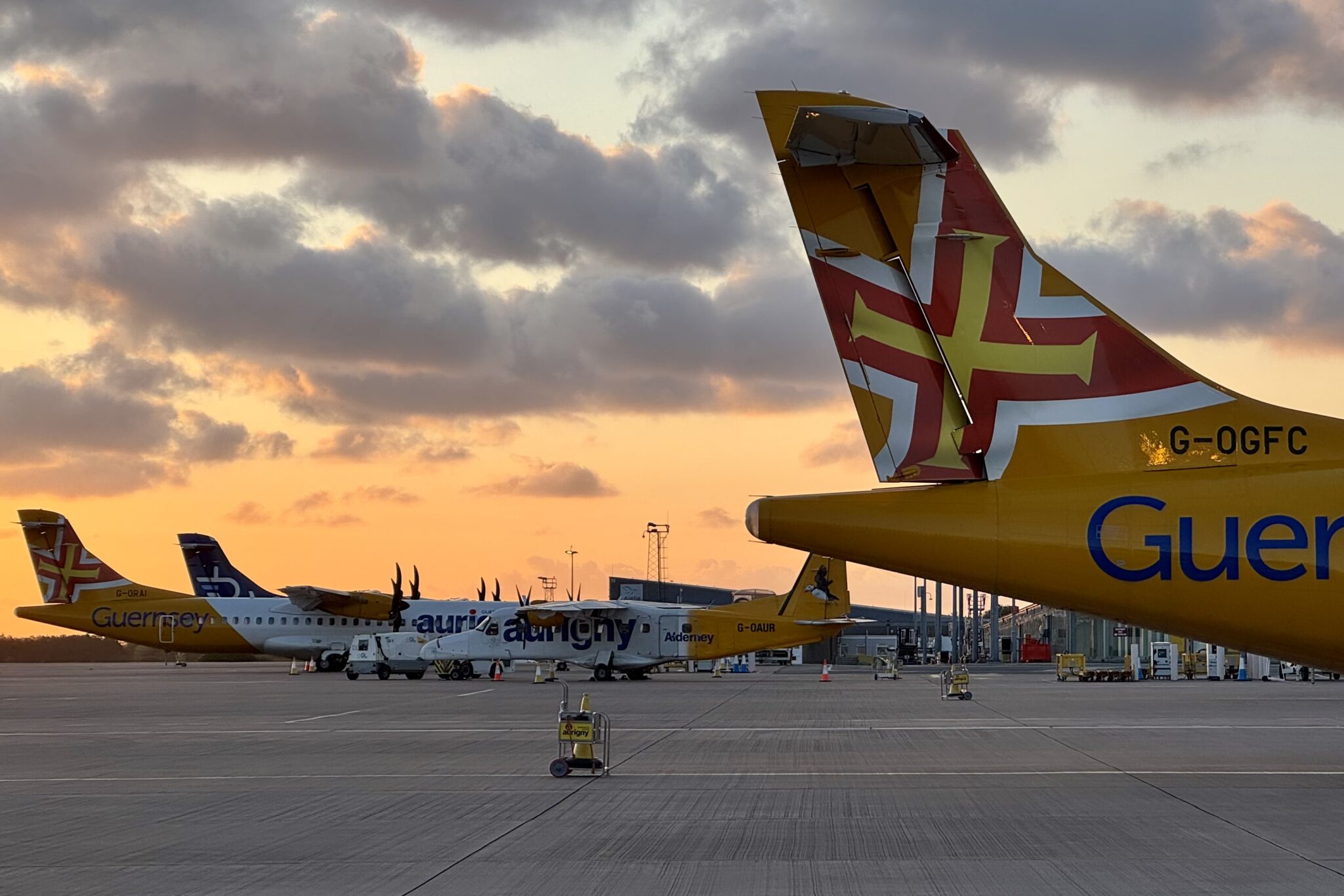 An image of the back of a Guernsey aircraft, yellow and red, amid cloudy and low sun skies, many other aircraft on the ground behind it.
