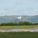 An aircraft landing in GLO airport, plane flying amid cloudy skies and near grassy terrain.