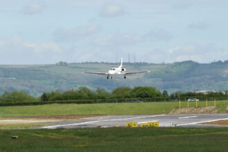 An aircraft landing in GLO airport, plane flying amid cloudy skies and near grassy terrain.