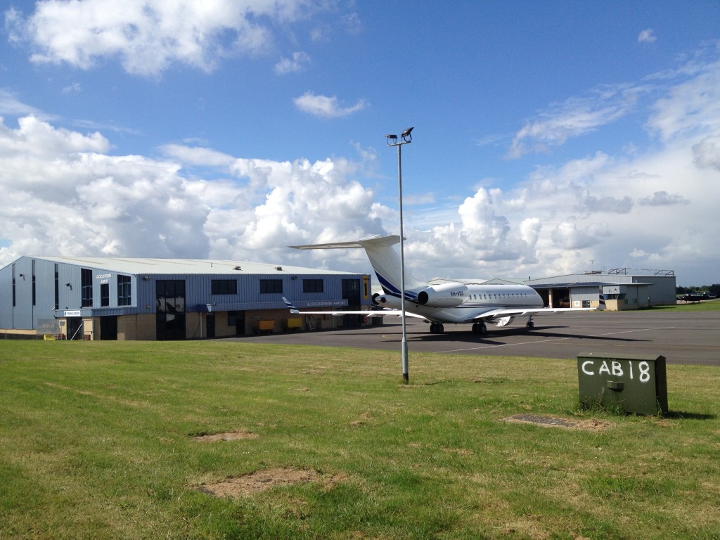 An image of GLO airport with blue skies and a few clouds, an aircraft on the ground and green grass beside it.