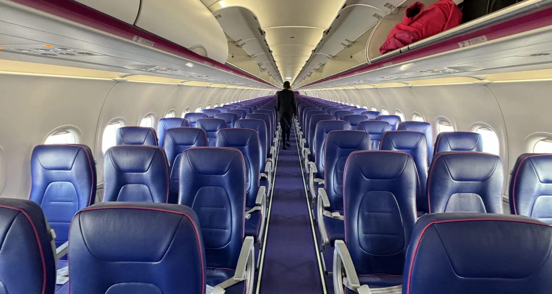 A Wizz Air aircraft interior, many blue seats facing the camera and overhead baggage lockers partially unlocked. A flight attendant stands towards the end of the image. 