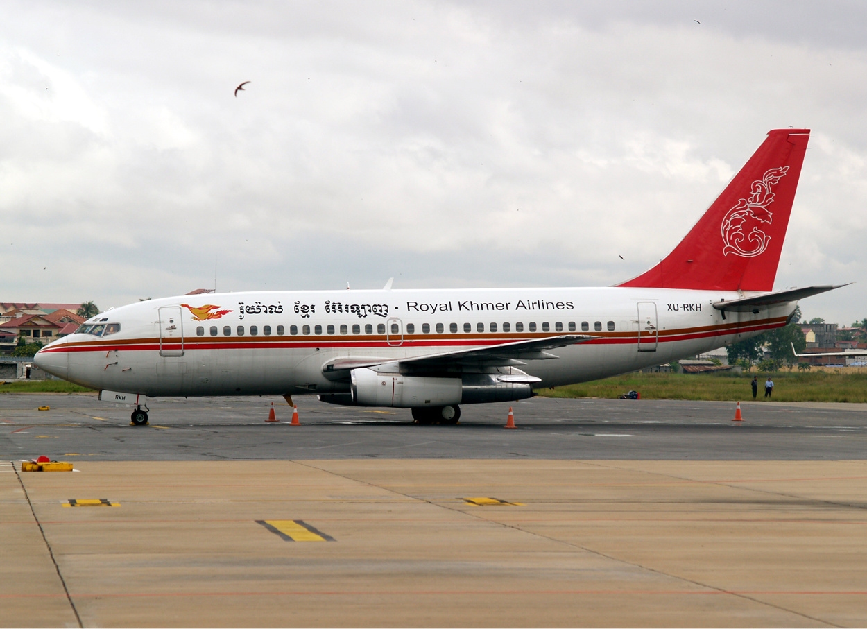 A shot of a Royal Khmer Airlines Boeing 737-200 parked in an airport at Phnom Penh International Airport. The aircraft is white with a red tail featuring a gold logo and a red and gold stripe running along the body.