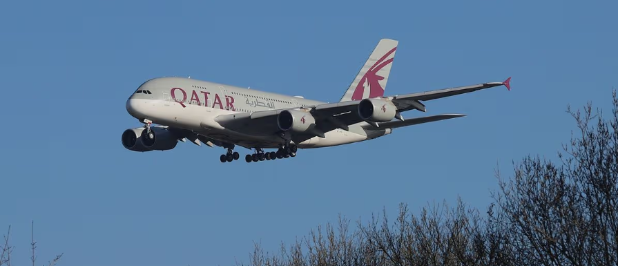 Qatar Airways Airbus A380 aircraft flying to the left side, blue sky, tree tops on the right.