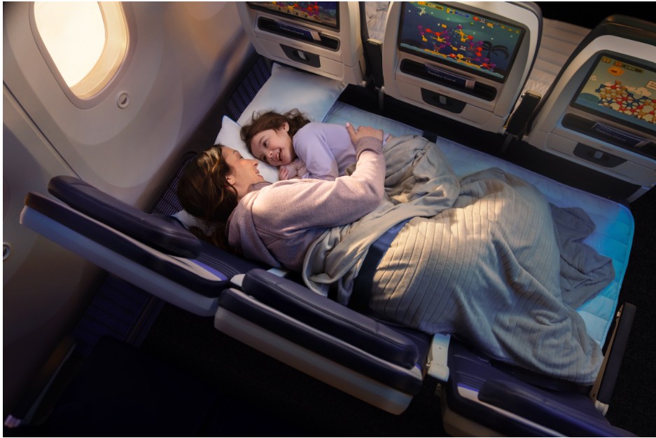 A woman and a young child lie side by side on fully reclined airplane seats converted into a flat bed, wrapped together under a light gray blanket.