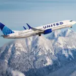 A United Airlines plane flying amid blue skies and a mountainous scenery below.
