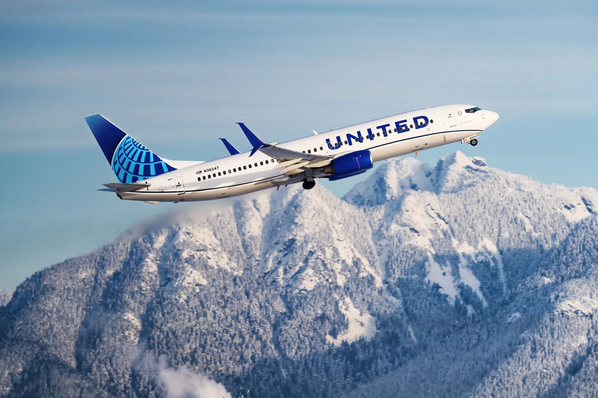 A United Airlines plane flying amid blue skies and a mountainous scenery below.
