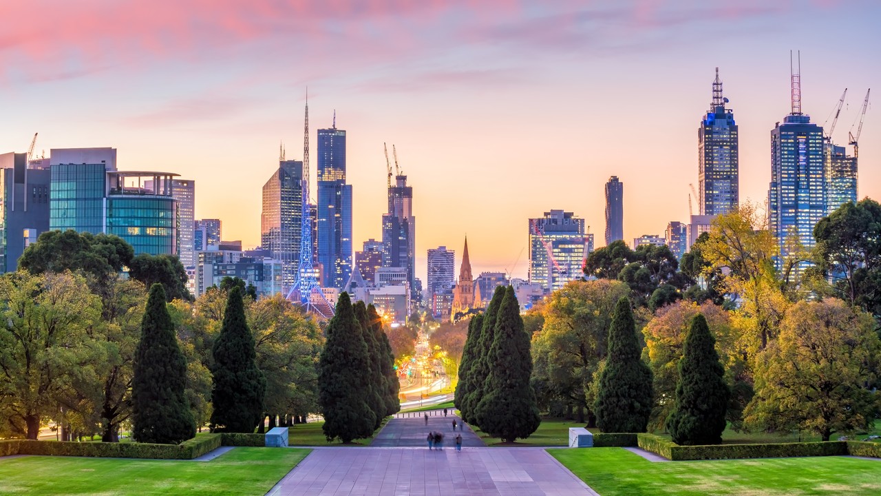 Image shows a view of Melbourne city skyline at twilight with park, trees and skyscrapers across a pinky, purple sky