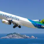 An Air Seychelles aircraft taking flight between blue, clear skies and blue seas, with an island in the centre of the sea below.