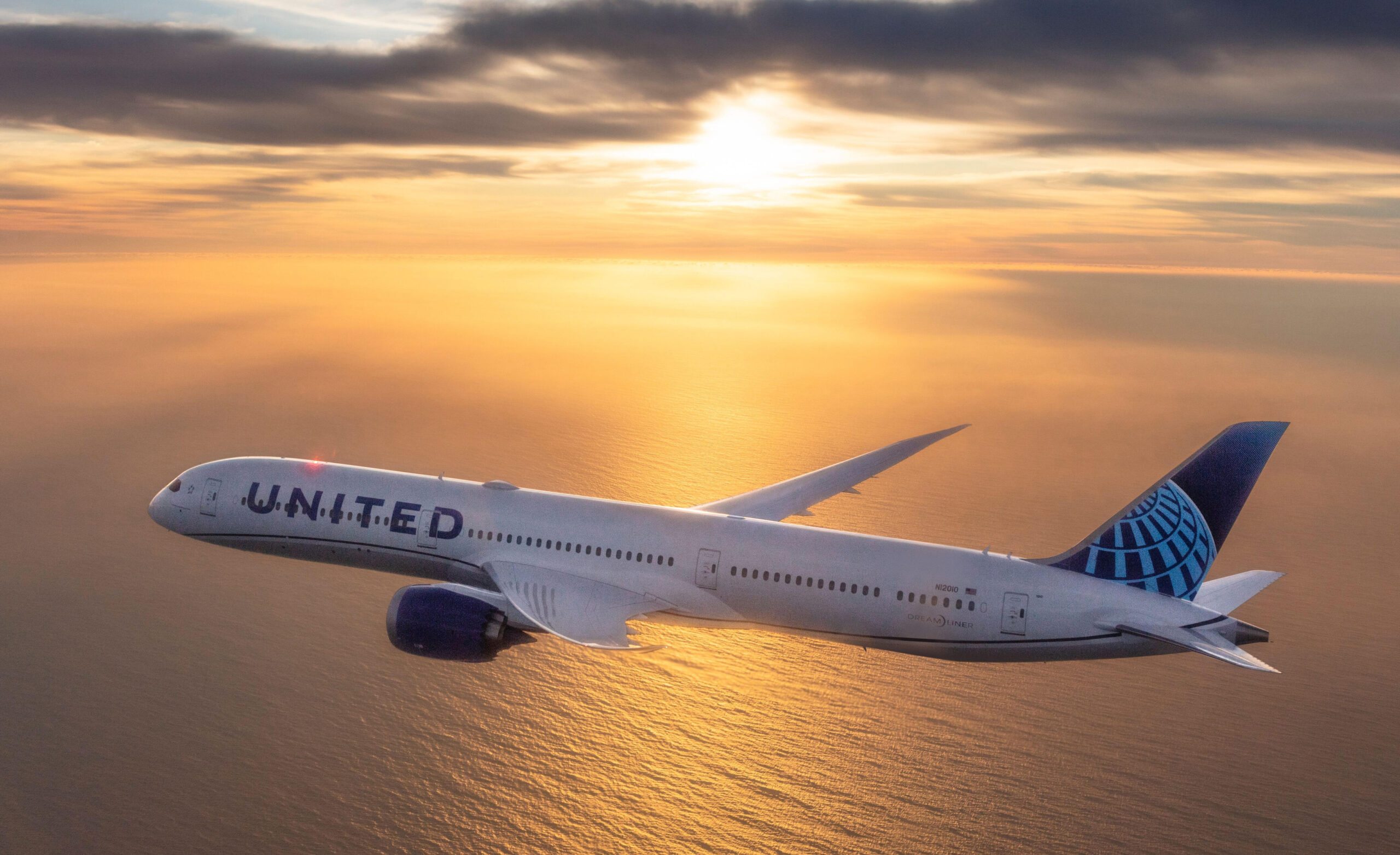 A United Airlines flight flying through a yellow and orange sunset across a body of water.