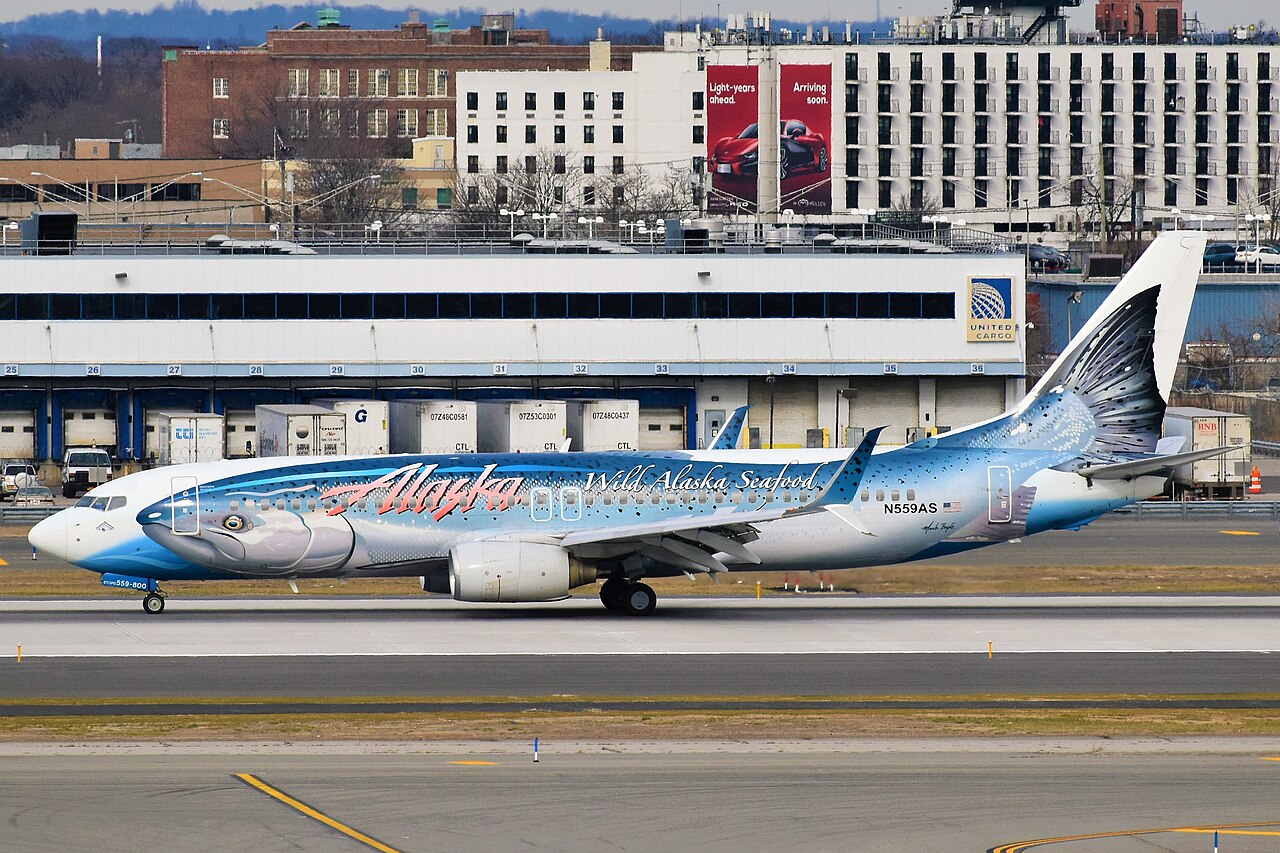 An Alaska Airlines jet with a distinctive salmon-themed livery taxis along a runway in front of airport cargo buildings.