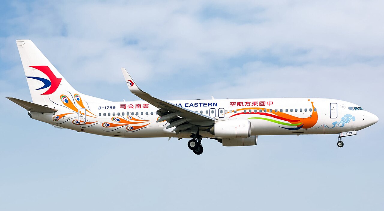 A China Eastern Airlines narrow-body airplane in a special peacock-themed livery flying against a partly cloudy sky, viewed from the side.