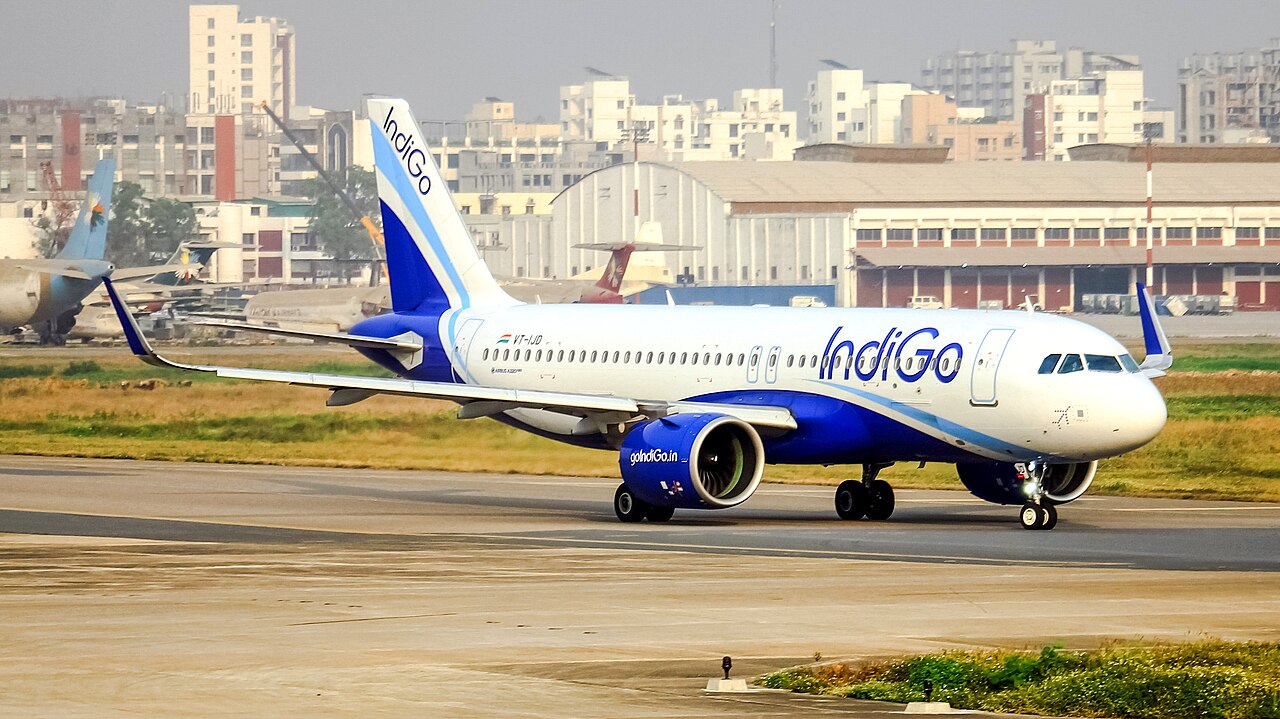 A white and blue IndiGo commercial airplane taxiing on a runway, viewed from the side in daylight.